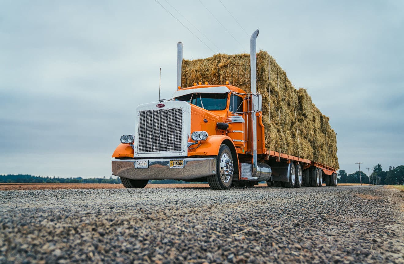 An orange Peterbilt on a gravel shoulder with a flatbed stacked and strapped full of hay bales